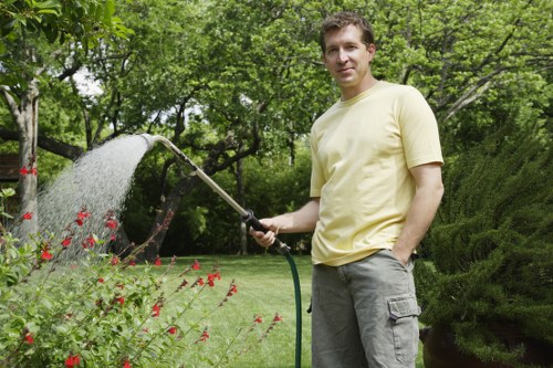 A man standing in a lush, well-maintained garden in Palmers Green, London, is watering a variety of flowering plants and shrubs using a garden hose. The garden features a neatly trimmed lawn with dense, vibrant green grass in the foreground, bordered by flower beds filled with red and pink flowers. In the background, there are tall trees with leafy canopies providing shade, and a neatly shaped hedge is visible on the right side. The lawn area is bordered by soil pathways or garden beds, creating a structured landscape. The weather appears mild and sunny, with bright natural light illuminating the scene. This outdoor space showcases typical residential garden features, reflecting professional gardening and lawn care practices that a service like Hedge Trimming Palmers Green might maintain for local homeowners, creating an inviting and well-kept garden environment.