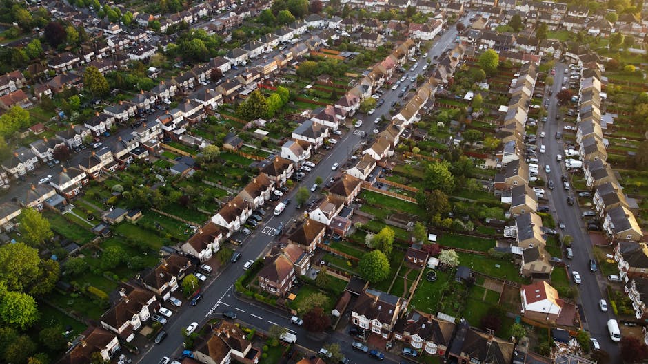 An aerial view of a residential suburb in Palmers Green N13 displaying neatly arranged houses with pitched roofs, lush green gardens, and tree-lined streets. The gardens feature a variety of textures 